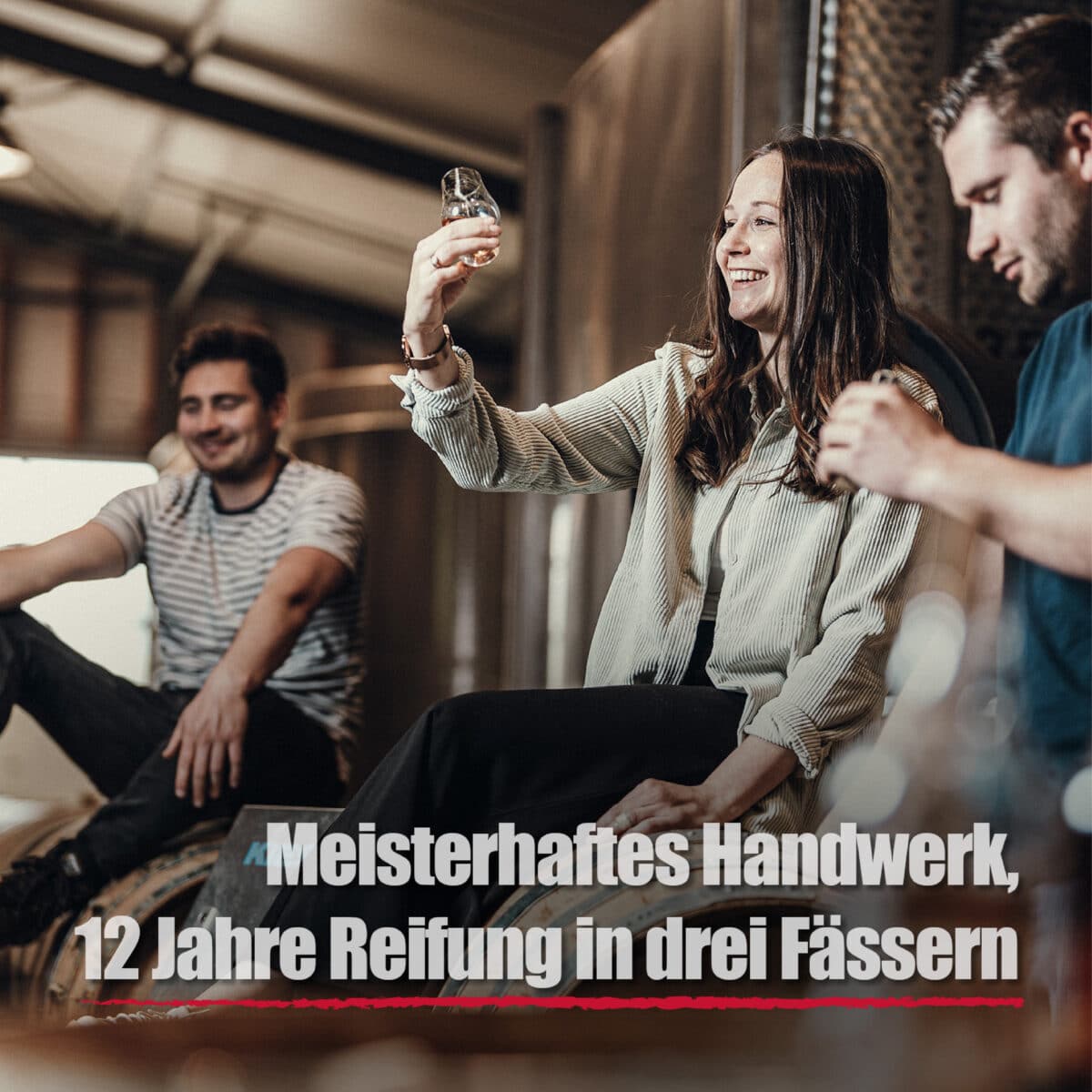 Drei Personen sitzen in einer Schnapsbrennerei zusammen, eine Frau hält ein Glas hoch und prüft dessen Inhalt. Der Text lautet: Black Wood Rum - meisterhaftes Handwerk, 12 Jahre gereift in drei Fässern.