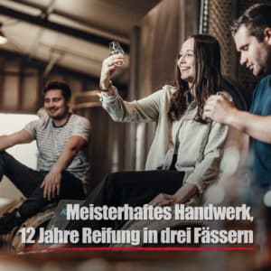 Drei Personen sitzen in einem Haus zusammen, eine Frau hält ein Glas in der Hand und prüft es. Der Text lautet: Black Wood Whisky - Meisterhaftes Handwerk, 12 Jahre Reifung in drei Fässern.