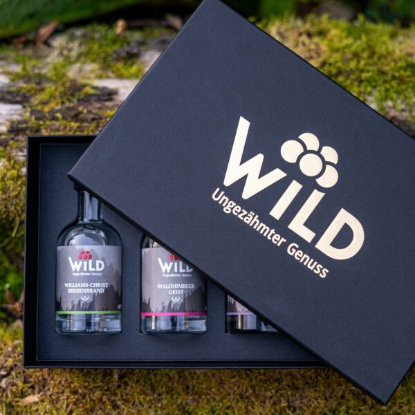 Ein schwarzer Geschenkkarton mit der Aufschrift Wild Tasting-Box Edelbrände & Geiste ist im Freien auf Moos teilweise geöffnet und zeigt drei kleine Flaschen mit Spirituosen.