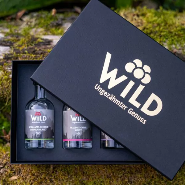 Ein schwarzer Geschenkkarton mit der Aufschrift Wild Tasting-Box Edelbrände & Geiste ist im Freien auf Moos teilweise geöffnet und zeigt drei kleine Flaschen mit Spirituosen.