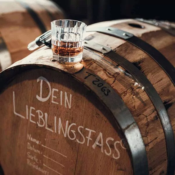 Ein Glas Whiskey steht auf einem Holzfass mit der Aufschrift Dein Lieblingsfass, auf dem ein Tropfer und ein handgeschriebener Text zu sehen sind.