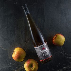 Eine Flasche Wildapfelwein liegt auf einer dunklen Schieferplatte neben drei roten und gelben Äpfeln.