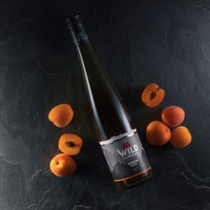 Auf einer dunklen Steinfläche liegt eine Flasche Wild Apricot Gold Wine, umgeben von mehreren ganzen und halbierten Aprikosen.