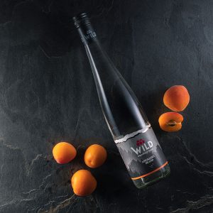 Eine Flasche Wild Apricot Brandy liegt auf einer dunklen Fläche, umgeben von vier ganzen Aprikosen und einer halbierten Aprikose.