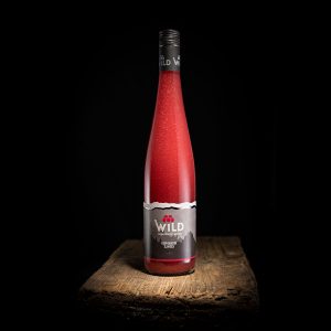 Eine rote Getränkeflasche mit der Aufschrift Wild steht aufrecht auf einer rustikalen Holzfläche vor einem schwarzen Hintergrund.