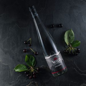 Eine Flasche Wildes Kirschwasser liegt auf einer dunklen Steinfläche, umgeben von Kirschen und grünen Blättern.