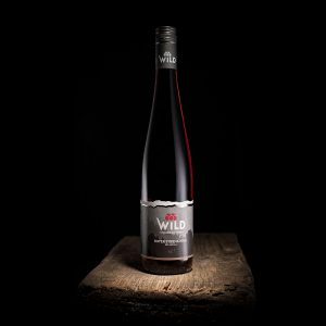 Eine Flasche Wild Rotweintrocken-Rotwein steht aufrecht auf einer rustikalen Holzfläche vor schwarzem Hintergrund.