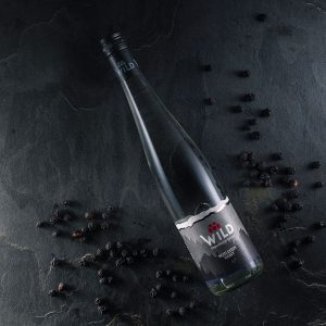 Eine durchsichtige Glasflasche mit der Aufschrift Wild Sour Cherry Fruit Guts liegt auf einer dunklen Steinfläche, umgeben von verstreuten getrockneten schwarzen Beeren.