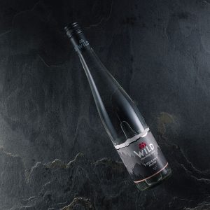 Eine Klarglasflasche mit der Aufschrift Wild liegt auf einer dunklen Steinoberfläche. Das Etikett zeigt ein minimalistisches Bergdesign mit roten Beeren.