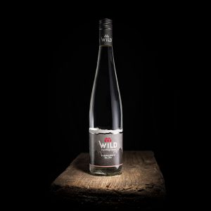 Eine Klarglasflasche mit Wild Topinamur Brandy mit schwarzem Etikett und Verschluss, platziert auf einer rustikalen Holzoberfläche vor einem schwarzen Hintergrund.