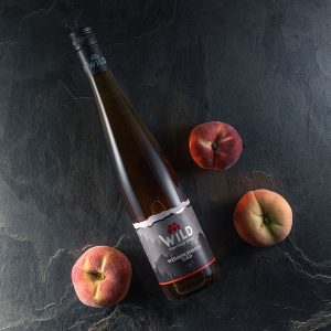 Eine Flasche Wilder Wein liegt auf einer dunklen Steinfläche, umgeben von drei flachen Pfirsichen.