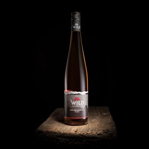 Eine Flasche Wilder Wein mit einem schwarz-silbernen Etikett steht auf einer hölzernen Oberfläche vor einem schwarzen Hintergrund.