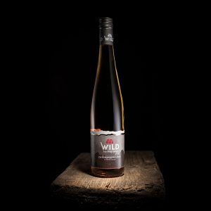 Eine Flasche Wilder Wein mit einem silbernen Etikett steht auf einer rustikalen Holzfläche vor einem schwarzen Hintergrund.
