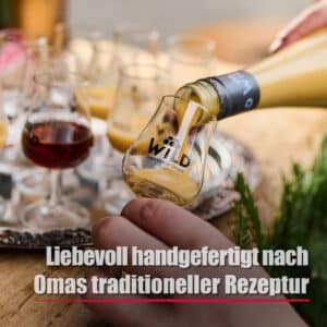 Eine Hand gießt Eierlikör Haselnuss, einen hellen Likör, aus einer Flasche in ein kleines Glas mit der Aufschrift "WILD", während im Hintergrund weitere gefüllte Gläser auf einem Tablett stehen und ein deutscher Text das Bild überlagert.