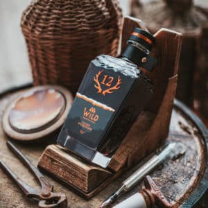 Eine Flasche Elixier Wildschlehe steht auf einem Holzständer auf einem Fass, umgeben von rustikalen Gegenständen und Weidenkörben.