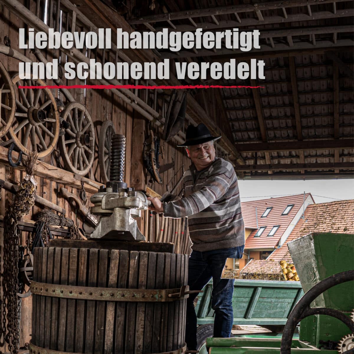 Ein Mann bedient eine traditionelle hölzerne Weinpresse in einer rustikalen Scheune mit alten Wagenrädern; der deutsche Text lautet: Liebevoll handgefertigt und sanft veredelt Schwarzer Johannisbeerlikör.