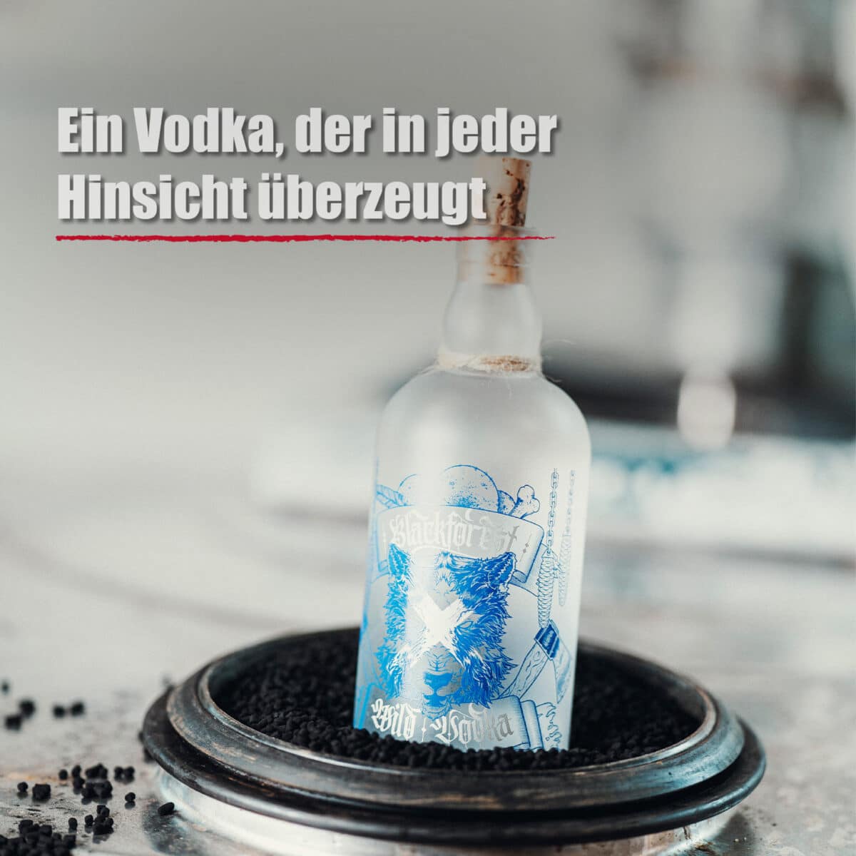 Eine Flasche Blackforest Wild Vodka mit Milchglas und blau-weißem Etikett steht aufrecht in einer runden Metallschale, die mit schwarzen Körnern gefüllt ist. Oben ist ein deutscher Text zu lesen.