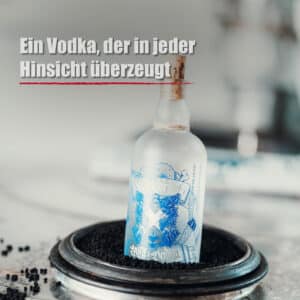 Eine Flasche Blackforest Wild Vodka mit Milchglas und blau-weißem Etikett steht aufrecht in einer runden Metallschale, die mit schwarzen Körnern gefüllt ist. Oben ist ein deutscher Text zu lesen.
