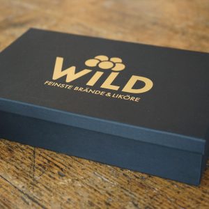 Eine schwarze rechteckige Box mit der Aufschrift "Wild Tasting-Box Goldbrände" und goldener Schrift steht auf einer Holzfläche.