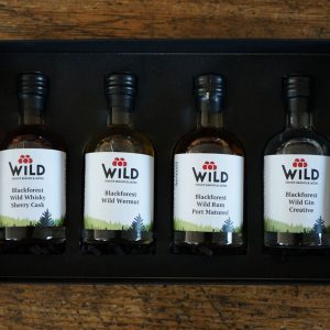 Die Wild Tasting-Box Goldbrände besteht aus vier kleinen Flaschen - Whisky, Wermut, Rum und Gin -, die hübsch in einer schwarzen Box auf einer hölzernen Oberfläche angeordnet sind.