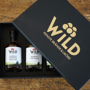 Die Wild Tasting-Box Goldbrände enthält drei Flaschen - Blackforest Wild Whisky Sherry Cask, Blackforest Wild Vermut und eine dritte, dazu passende Flasche -, die elegant in einer Geschenkbox präsentiert und auf einer Holzfläche präsentiert werden.