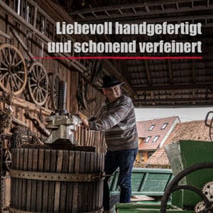 Ein älterer Mann keltert Wein in einer rustikalen Scheune; darüber steht auf Deutsch "Liebevoll handgefertigt und schonend verfeinert", was sich auf Wurzelfeuer bezieht.