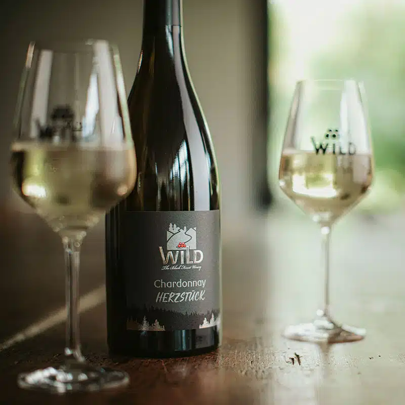 Eine Flasche Wild Chardonnay Herzstück wird auf einen Holztisch zwischen zwei gefüllte Weingläser gestellt.