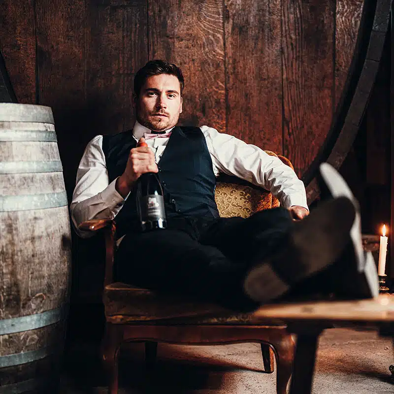 Wild-Weine_Unterseite-Bild-3 Ein Mann in formeller Kleidung sitzt auf einem alten Stuhl, legt die Fรผรe hoch und hรคlt eine Flasche Wein in der Hand. In der Nรคhe stehen Holzfรคsser und eine brennende Kerze, was auf eine rustikale Umgebung hindeutet.