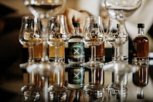 Eine Hand greift nach einer Online-Verkostungsbox inmitten mehrerer Whiskey-Gläser, die zur Verkostung bereitstehen, wobei die Schnapsflaschen auf einer reflektierenden Oberfläche ausgestellt sind.