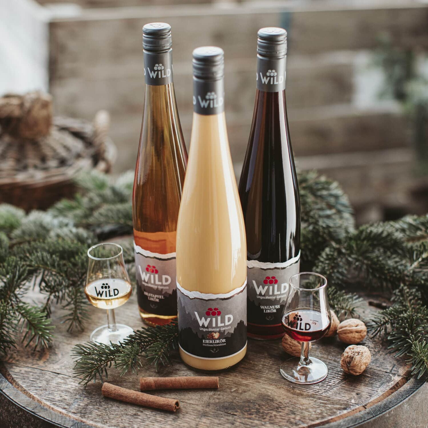 Drei Weinflaschen der Marke Wild und zwei Gläser sind auf einer hölzernen Fläche mit Tannenzweigen, Walnüssen und Zimtstangen als Dekoration angeordnet.