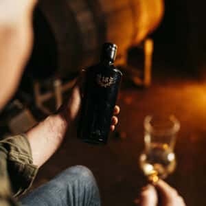 Eine Person hält eine schwarze Flasche 1986 Wilde Schlehe aus dem Kastanienholzfass in einer Hand und ein Glas hellen Schnapses in der anderen, mit einem unscharfen Fass im Hintergrund.