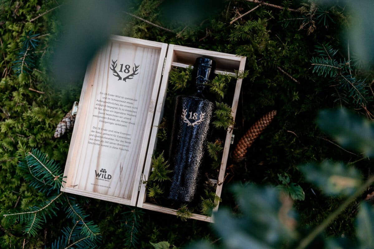 Eine schwarze Flasche mit der Aufschrift 1986 Wilde Schlehe aus dem Kastanienholzfass steht in einer offenen Holzkiste mit Text, umgeben von Tannenzweigen und -zapfen auf einem moosbewachsenen Boden.