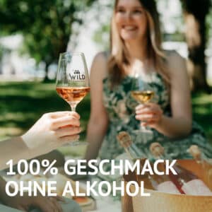 Zwei Frauen genießen LÚVO Hibiskus x Basilikum - Alkoholfrei im Freien und nippen aus Weingläsern mit Flaschen auf einem Picknicktisch. Text: 100% Geschmack ohne Alkohol.