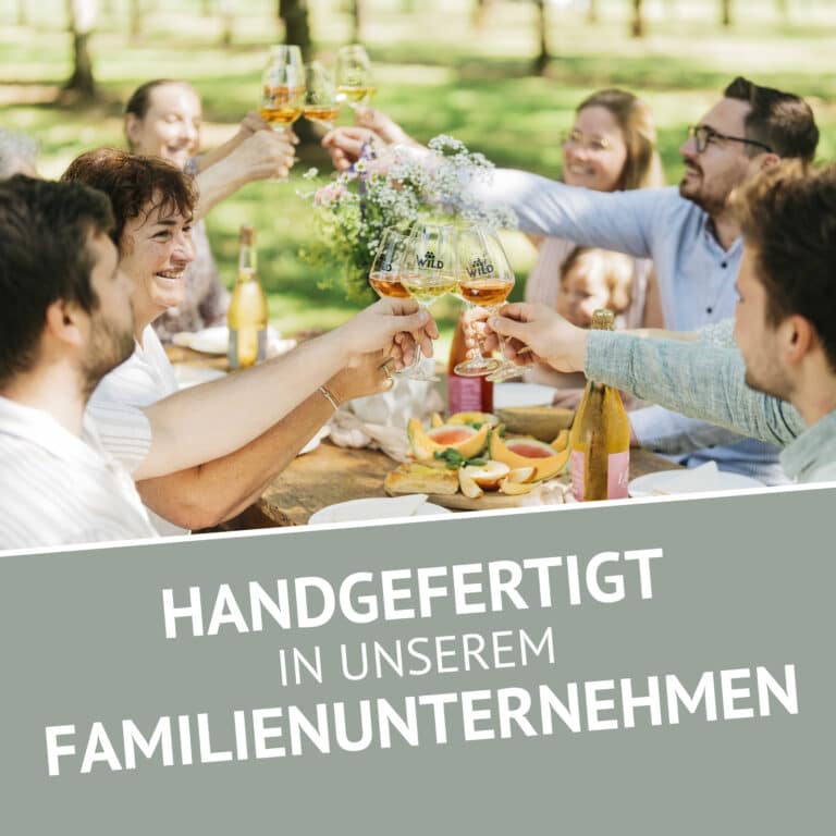 Eine Gruppe von Personen genießt eine Mahlzeit im Freien und stößt mit Gläsern von LÚVO Hibiskus x Basilikum - Alkoholfrei an, wobei am unteren Rand der Text Handgefertigt in unserem Familienunternehmen erscheint.
