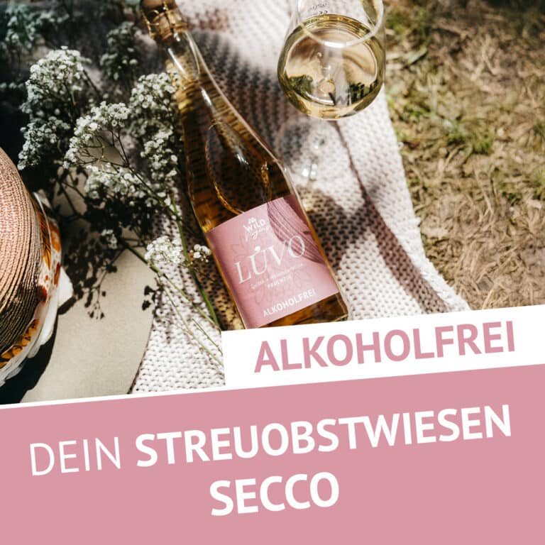 Eine Flasche LÚVO Quitte x Holunderblüte - Alkoholfrei ruht auf einer Decke neben einem Weinglas und Blumen. Der Text lautet: ALKOHOLFREI DEIN STREUOBSTWIESEN SECCO.