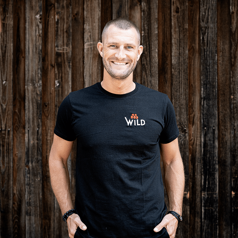 Ein Mann mit kurzen Haaren und gestutztem Bart steht vor einer Holzwand, lächelt und trägt ein schwarzes T-Shirt mit dem Aufdruck WILD.