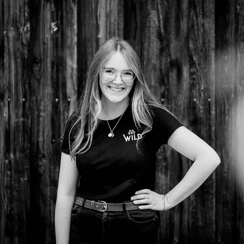 Eine Frau mit langen Haaren und Brille steht lächelnd mit einer Hand an der Hüfte und einem WILD-T-Shirt vor einer Holzwand.