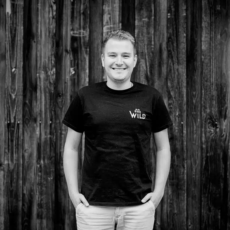Eine lächelnde Person mit einem WiLD-T-Shirt steht mit den Händen in den Taschen vor einer Holzbohlenwand.
